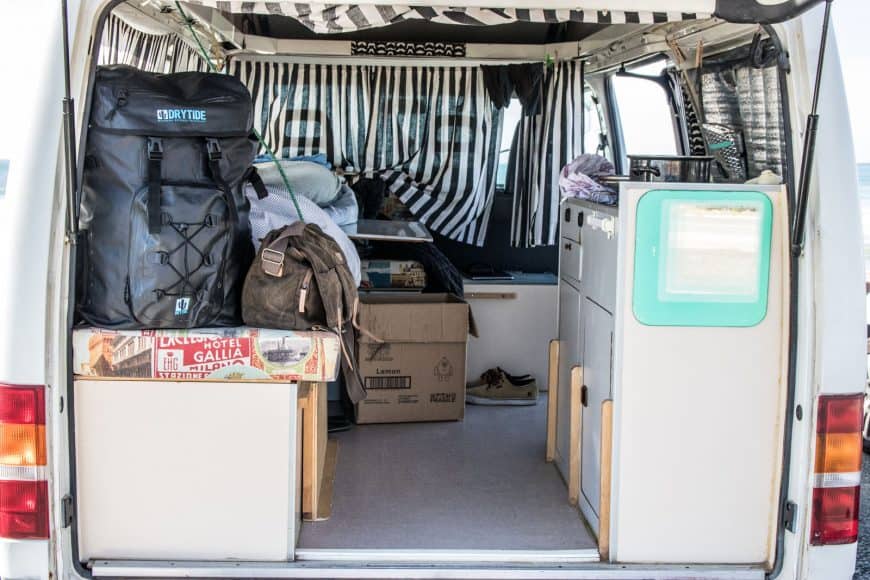 A look into our van from the back. There is even a mirror mounted on the side of the kitchen cabinet. DryTide waterproof backpack in the left is awesome to protect all your stuff from sand and water and it folds down next to nothing when you are not using it so it's a perfect backpack for van life.